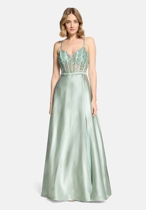 Frau mit blondem Haar, die ein hellgrünes Satinkleid mit besetztem, transparentem Oberteil und Spaghettiträgern trägt, steht vor einem schlichten Hintergrund.