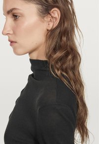 Black turtleneck sweater, fitted design, soft texture, and ribbed accents. Hair styled in loose waves cascading over the shoulder.