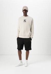 Lys beige teksturert hettegenser med NY-logo, kombinert med svarte mesh-shorts og hvite joggesko, komplettert med en lysebrun caps.