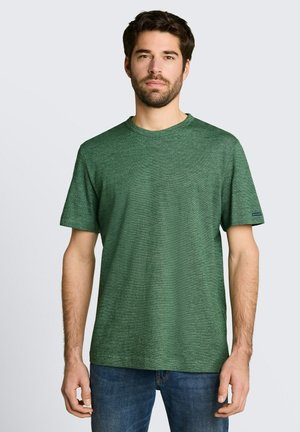 Man met kort bruin haar en baard, die een effen groen T-shirt met korte mouwen en blauwe spijkerbroek draagt, staand tegen een lichte achtergrond.