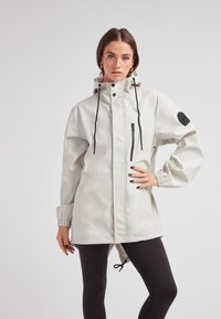 Lichtgrijze waterdichte jas met een capuchon, verstelbare trekkoorden, een frontaal rits en zijzakken. Voorzien van elastische manchetten en een logo patch.