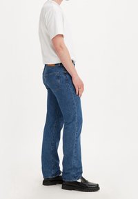 Blue denim jeans with a straight cut, high waist, and five pockets, worn with a plain white t-shirt and black slip-on shoes.