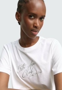 Weißes Baumwoll-T-Shirt mit gesticktem Text "5th Ave Club" in metallischem Silber. Klassischer Rundhalsausschnitt und kurze Ärmel. Glatte Textur.