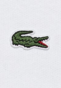 Logotipo de cocodrilo bordado en verde sobre un fondo de tela blanca, con detalles texturizados y un acento rojo en la boca.