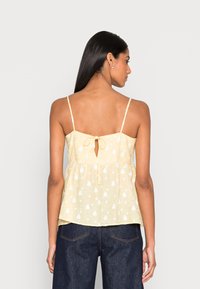 Top amarelo sem mangas com padrões de gota brancos, detalhe de laço nas costas e design peplum. Tecido leve com uma textura suave.