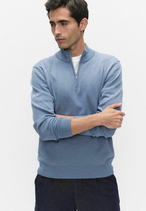 Mies sinisessä puolivetoketjuneuleessa ja tummissa housuissa seisoo käsivarret ristissä ja katsoo vasemmalleen yksinkertaisen valkoisen taustan edessä.