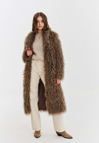 Dlouhý, chlupatý hnědý kabát z umělé kožešiny přes béžový svetr a krémové kalhoty. Kabát má volný střih a tmavý podšívku. Na nohou jsou nošeny špičaté hnědé boty.