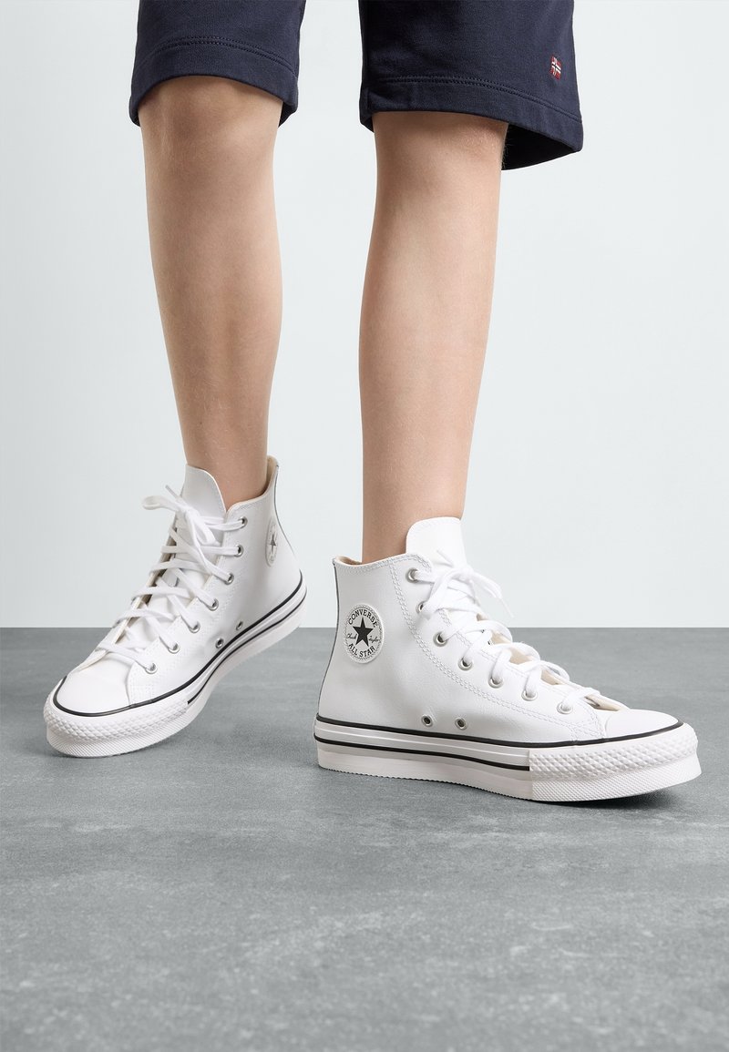 Enfant portant des baskets montantes blanches Converse et un short bleu marine, debout sur un sol gris devant un fond uni.