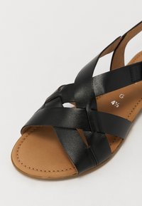 Black leather sandal with crisscross straps, open toe, and a tan sole. Features smooth texture and stitching details on the sole.