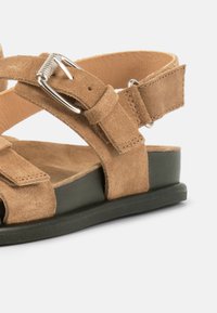 Καφέ σανδάλι από σουέτ με ρυθμιζόμενο λουράκι και ασημένια αγκράφα, με μαύρη σόλα σε λευκό φόντο.