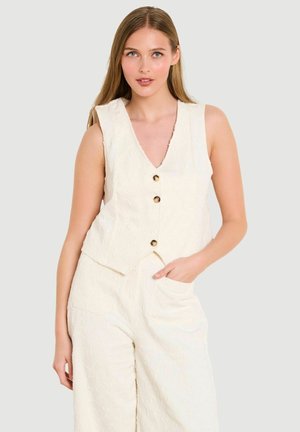 Młoda kobieta ubrana w kremową bezrękawnikową kamizelkę o fakturze, zapinaną na trzy guziki, oraz dopasowane spodnie z wysokim stanem, z jedną ręką w kieszeni, na neutralnym tle.