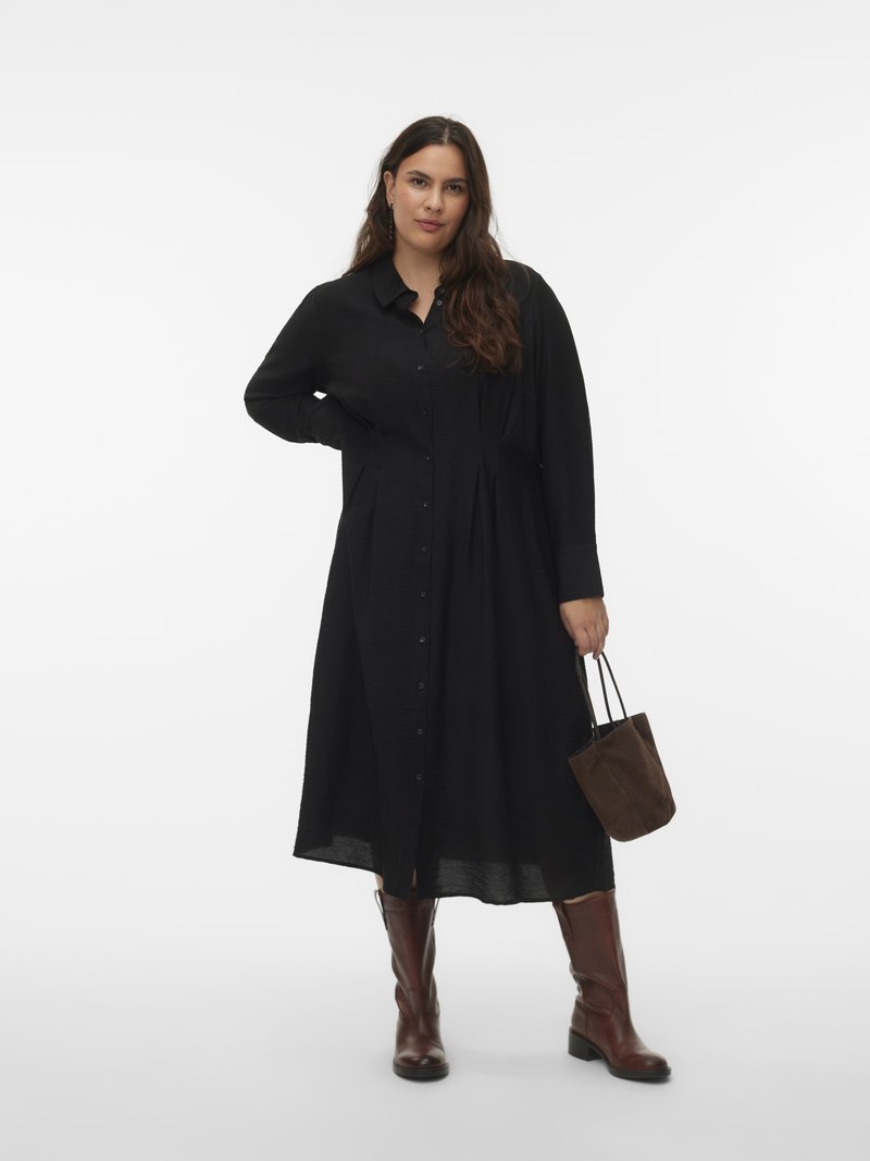 Ženska, oblečena v dolgo črno obleko z gumbi, rjavi usnjeni škornji, drži majhno rjavo semišasto torbico na belem ozadju.