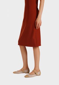 Ballerine in rete beige con punta cappuccino e cinturino alla caviglia, abbinate a un vestito fitted color ruggine che mette in risalto una silhouette slanciata.