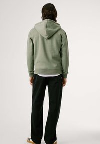 Hombre con sudadera verde, pantalones negros y zapatillas blancas, de pie de espaldas contra un fondo blanco liso.