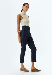 Crème kleurige geribbelde mouwloze coltrui, high-waisted marineblauwe broeken en bruine textuur slip-ons met een puntige neus.