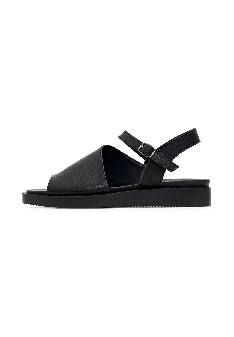 Sandelă neagră cu superior din piele, vârful deschis și curea de tip slingback. Prezintă o talpă plată și o cataramă ajustabilă pentru o fixare sigură.