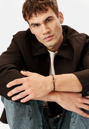 Schwarze Jacke mit Reißverschluss, weißes Hemd darunter und blaue Jeans aus Denim. Ein schwarzes Perlenarmband mit goldenem Akzent am Handgelenk.