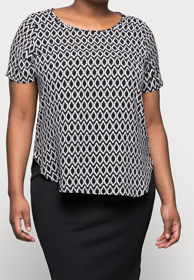 Femme portant un haut à manches courtes à motifs noir et blanc et une jupe noire ajustée, debout devant un fond uni.