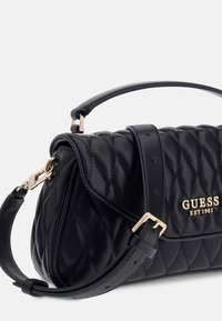 Borsa Guess in pelle trapuntata nera con logo dorato, tracolla regolabile e manico superiore su uno sfondo grigio chiaro.
