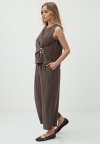 Bruine mouwloze top met een vooraan draai-detail en ringaccent, gecombineerd met loszittende bruine broek en platte geweven schoenen.