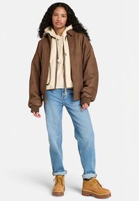 Bruine bomberjas van nep leer met geribbelde manchetten, gedragen over een beige hoodie en gecombineerd met lichtblauwe jeans en beige laarzen.