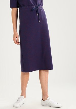 Femme portant une jupe midi bleu marine à taille coulissante, associée à des baskets blanches décontractées, debout devant un fond clair.