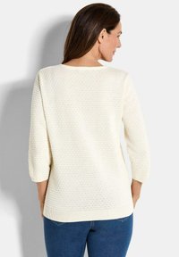 Frau in einem strukturierten weißen Langarmpullover und blauen Jeans, von hinten vor einem schlichten hellen Hintergrund abgebildet.