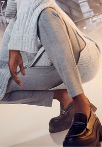 Černé platformové mokasíny s tlustými gumovými podrážkami, s prošívaným designem a kovovým akcentem. Modelka nosí šedé zkrácené kalhoty a svetr.