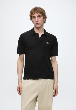 Noor mees helepruunide lokkis juustega kannab musta tekstuuriga lühikeste varrukatega polosärki ja beeže pükse, seisab valgel ühevärvilisel taustal.