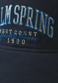 Casquette marine avec le texte brodé "PALM SPRING" en bleu et crème, présentant un visière structuré et un tissu texturé. Détails dorés inclus.
