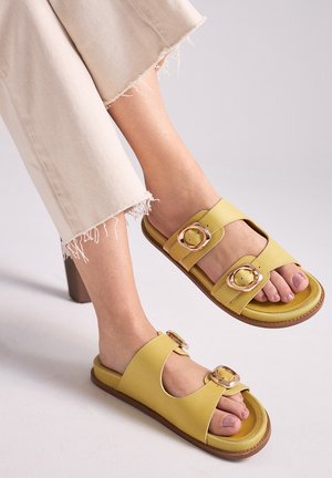 Pies con sandalias de doble hebilla color amarillo claro combinadas con pantalones cortos beige claro con dobladillos deshilachados sobre un fondo gris claro.