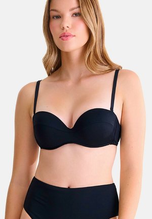 Vrouw draagt een zwarte strapless beha en hoogwaterondergoed, staand tegen een effen lichte achtergrond met een neutrale gezichtsuitdrukking.