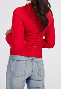 Veste matelassée rouge à manches longues, avec une coupe ajustée et un petit logo, associée à un jean bleu clair avec des poches arrière.
