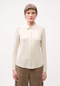 Langärmlige cremefarbene Bluse mit Kragen und durchgehender Knopfleiste. Glatte Textur, figurbetontes Design und subtile Nahtdetails.