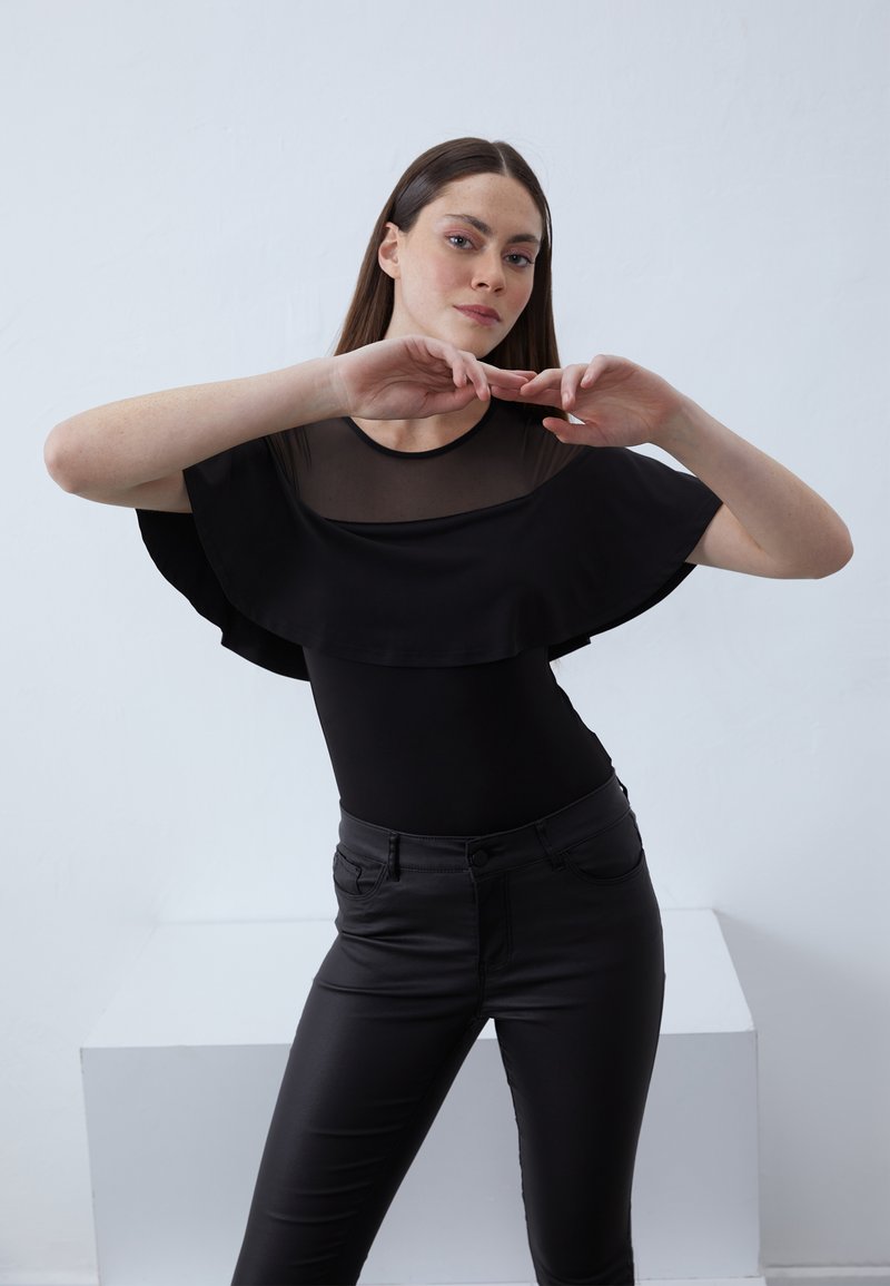 Musta paita, jossa on läpinäkyvä päällyskerros, leveällä frillahelmalla ja mukautuvalla bodicella. Yhdistetty hoikilla mustilla slim-fit-housuilla.