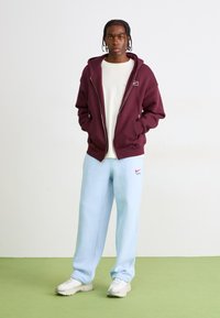 Bordeaux zip-up hoodie over en hvid skjorte, parret med lyseblå sweatpants med et rødt Nike-logo og hvide sneakers.