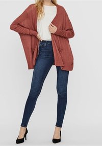 Cardigan i rustfarve, let strik, åben front med to lommer, kombineret med mørke skinny jeans og sorte hæle. Glat tekstur, afslappet pasform.