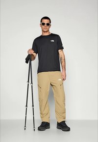 Bărbat purtând ochelari de soare, tricou negru, pantaloni cargo bej și bocanci de drumeție, ținând două bâte de trekking negre, pe un fundal simplu.