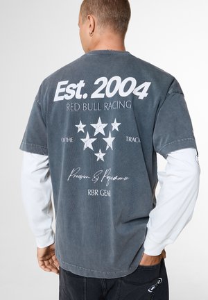 Person trägt ein verblasstes schwarzes Red Bull Racing T-Shirt mit weißem Text und Sternen, darüber ein weißes Langarmshirt.