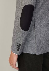 Chaqueta ajustada con patrón de espiga. Tejido gris con parches negros en los codos. Tres botones negros en los puños. Textura suave, corte estructurado.