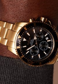 Goldfarbene Edelstahl-Uhr mit schwarzem Zifferblatt, drei Subdials und leuchtenden Markierungen. Verfügt über eine strukturierte Lünette und goldene Armbandglieder.