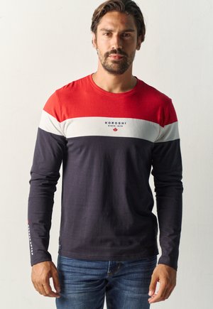 Hombre con barba que lleva una camisa de manga larga con bloques de color en rojo, blanco y azul marino con el texto "Koroshi" de pie contra un fondo claro y liso.