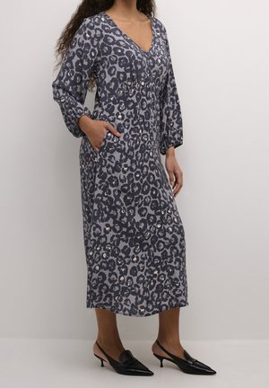 Robe à manches longues et col en V en tissu gris avec un imprimé léopard noir et beige clair, poches latérales et silhouette fluide. Associée à des talons noirs.