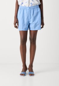 Shorts en coton bleu clair avec une ceinture élastique et un bouton à l'avant. Portés avec des sandales bleues à talons carrés et bouts ouverts. Texture lisse.