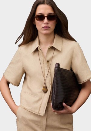 Kvinde iført beige kortærmet blazer og bukser, brune solbriller, langt vedhængsvedhæng, der holder en stor mørk tekstureret clutchtaske.