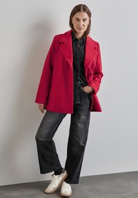 Abrigo de mezcla de lana rojo con solapa, combinado con una camisa negra de botones y jeans gris oscuro. Zapatillas blancas completan el atuendo.
