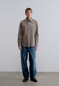 Καφέ πουκάμισο μακρυμάνικο με κουμπιά στο μπροστινό μέρος και γιακά, συνδυασμένο με μπλε τζιν τζιν, και μαύρα παπούτσια, στέκεται μπροστά σε απλό φόντο.