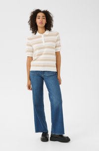 Polo rayé crème et beige, à manches courtes, avec trois boutons au col. Associé à un jean en denim bleu à jambes larges et des chaussures noires.