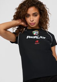 Vrouw met krullend haar, gekleed in een zwart Replay FC sportshirt met logo's, poseert met één arm gebogen achter haar hoofd tegen een effen achtergrond.