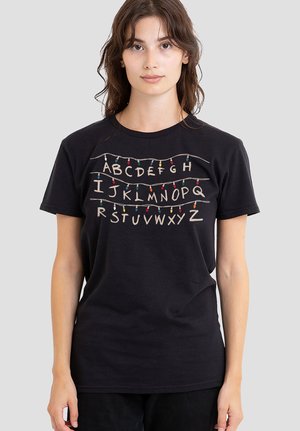 Junge Frau trägt ein schwarzes T-Shirt mit den Buchstaben des Alphabets von A bis Z, die durch farbige Lichterketten über drei Zeilen verbunden sind.